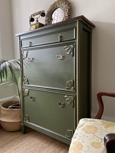 a green dresser sitting in front of a window next to a chair and potted plant