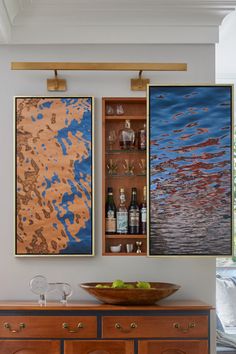 two framed art pieces on the wall above a wooden cabinet with wine glasses in it