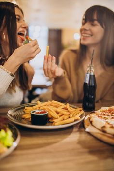 two women sitting at a table eating pizza and french fries with ketchup on the side