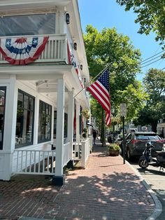 an american flag hanging from the side of a white building on a brick sidewalk next to parked cars