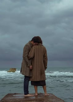 two people standing on the edge of a pier looking out at the ocean under a cloudy sky