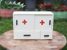 a white cabinet with red crosses painted on it sitting on a wooden table in front of some potted plants