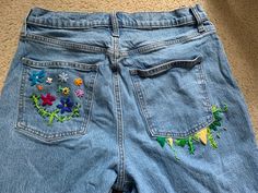 a pair of blue jeans with embroidered flowers on the front and back pockets, sitting on a carpeted floor