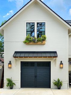 a white house with two black garage doors and plants in the window boxes on each side