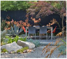 a table and chairs in a garden with rocks, gravel and plants around it is surrounded by trees