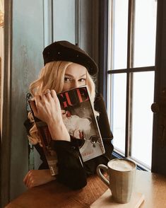 a woman sitting at a table with a coffee cup and magazine on her lap, next to a window