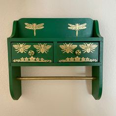 a green and gold painted wooden shelf with two bees on the top, one drawer open