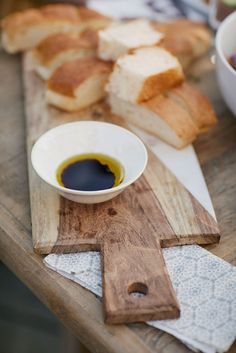 bread and olive oil on a cutting board next to bowl of salad dressing, with other food items in the background