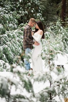 a pregnant woman standing next to a man in the snow surrounded by pine trees and evergreens