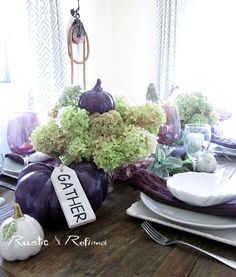 the table is set for thanksgiving dinner with purple pumpkins and greenery on it