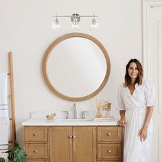 a woman standing in front of a bathroom vanity with a round mirror above the sink