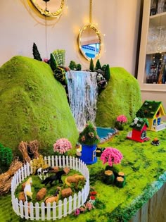 a table topped with fake grass covered in miniature houses and animals next to a waterfall