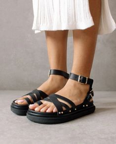 Person wearing ALOHAS BUCKLE UP SANDAL in black, featuring strappy espadrille platform design with ankle straps and silver buckles. The sandals have a thick sole accented by small studs near the straps. They are standing on a grey floor, and their lower legs are visible, wearing a white, ribbed knee-length skirt. Leather Gladiator Sandals, Sustainable Leather, Beautiful Sandals, Espadrilles Platform, Sandal Shoes, Buckle Sandals, School Project, Ankle Straps, Toe Rings