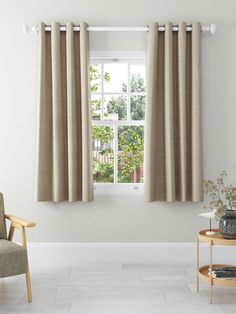 a living room with a chair, table and two windows covered in beige drapes