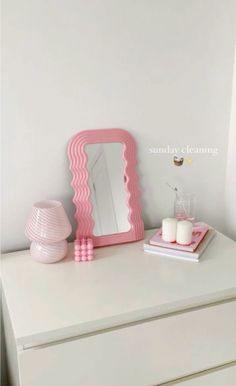 a white dresser topped with a pink mirror next to a candle and vase on top of it