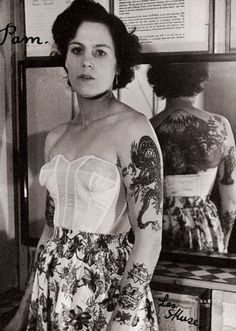 an old photo of a woman with tattoos on her arm and shoulder, standing in front of a mirror