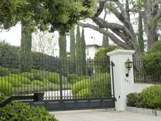 an iron gate with a clock on it in front of some trees and bushes near a house