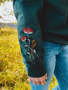 a person wearing a green sweater with red flowers embroidered on the sleeves and arm, standing in grass