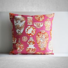 a pink pillow with gold and white vases on the front, sitting on a shelf