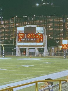 the scoreboard is lit up at night on an empty football field with people sitting in chairs