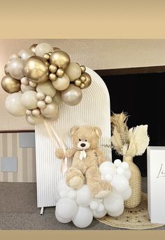 a teddy bear sitting on top of a basket filled with white and gold balloons in front of a fireplace