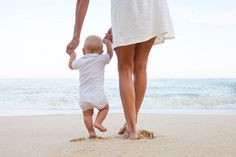 a woman holding the hand of a baby walking on top of a sandy beach next to the ocean