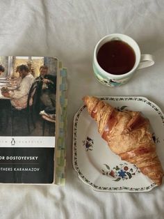 a croissant on a plate next to a coffee cup and book about food