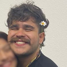 a man with a flower in his hair smiles at the camera while another man looks on