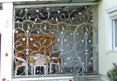 an iron gate with chairs on it in front of a house that is painted white