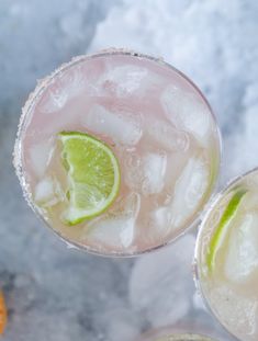 two glasses filled with ice and limes on top of a marble table next to an orange slice