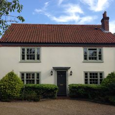 a white house with red tiled roof and two windows on each side, surrounded by hedges