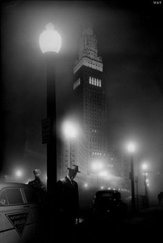 a black and white photo of a man standing next to a car at night in front of a tall building