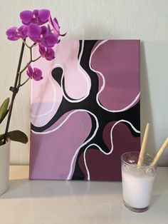 a vase with purple flowers sitting next to a painting on a white surface and a glass filled with milk