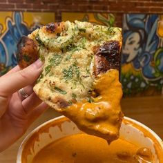 a person holding up a slice of pizza in front of a bowl of tomato soup