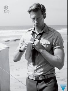 black and white photograph of a man tying his necktie on the beach by the ocean