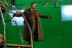 a man standing on top of a wooden chair in front of a green screen with his arms out