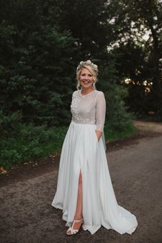 a woman in a white dress standing on a road with trees and bushes behind her