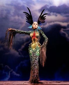 a woman dressed in an elaborate dress with wings on her head and arms outstretched, standing against a cloudy sky