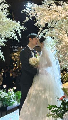 a bride and groom kissing in front of an outdoor wedding ceremony with white flowers on the trees