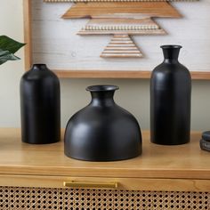 three black vases sitting on top of a wooden table next to a wall hanging