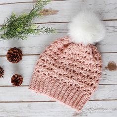 a crocheted hat with a white pom - pom sits next to pine cones