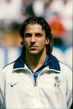 a man with long hair wearing a white jacket and blue collared shirt standing in front of a soccer field