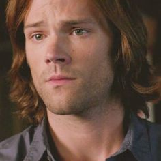 a man with long hair and blue eyes looks at the camera while wearing a black shirt