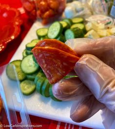 a person in white gloves holding up a piece of food on a cutting board with cucumbers and other vegetables