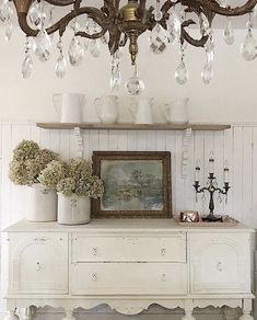 a chandelier hanging from the ceiling above a white dresser with vases and flowers on it