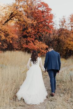 a bride and groom walking down a path in an autumn field with trees behind them