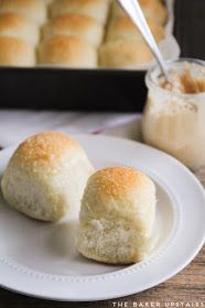 two rolls on a white plate next to a baking pan with bread in the background