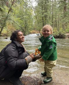 a woman kneeling down next to a little boy on a river bank with trees in the background