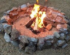 a fire pit made out of rocks and logs with flames coming from the top, on gravel ground