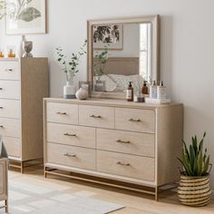 a bedroom scene with focus on the dresser and bed, mirror and plant in the corner